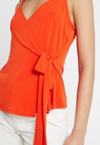 Top sans manches orange vif avec un nœud sur le côté, porté avec un pantalon blanc, montrant la taille et le bras sur un fond uni.