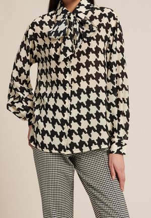 Femme portant un chemisier oversized pied-de-poule noir et blanc avec col à nouer en noeud papillon et un pantalon vichy noir et blanc, main posée sur la hanche.