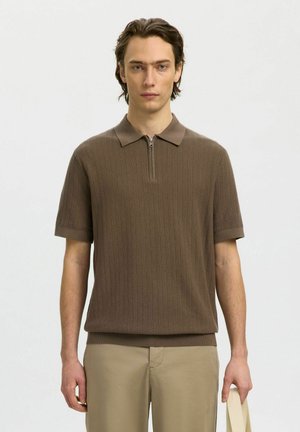 Jeune homme portant un polo en maille à manches courtes marron avec fermeture éclair, pantalon beige, tenant un sac fourre-tout de couleur claire devant un fond uni.
