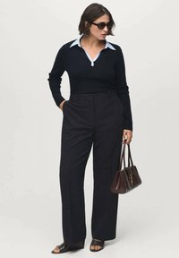 Woman wearing black long-sleeve top over light blue collared shirt, black wide-leg pants, black open-toe shoes, dark sunglasses, holding brown handbag.