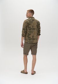 Kamuflāžas raksta, olīvu zaļš krekls ar pogām un uzrullētām piedurknēm, apvienots ar uzrullētām khaki šortiem un bēšām sandālēm. Novietots no aizmugures.