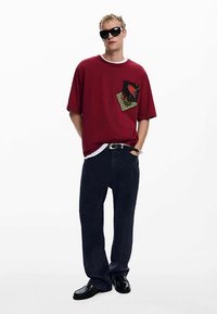 Tricou roșu supradimensionat cu un detaliu grafic pe buzunar, asortat cu pantaloni largi bleumarin închis și pantofi negri. Accesoriile includ ochelari de soare închiși la culoare.