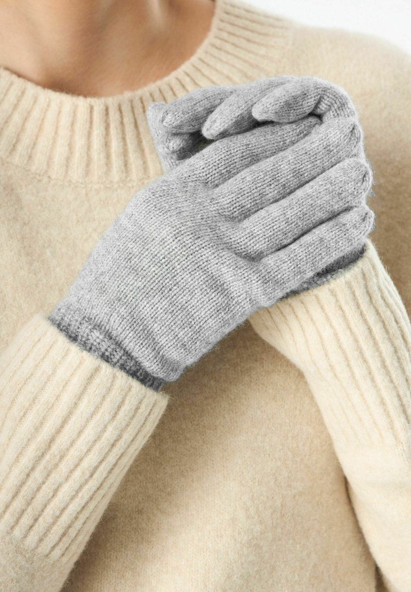 Gants tricotés gris avec des poignets côtelés, associés à un pull en tricot beige clair. Les gants présentent une texture douce et un subtil contraste de couleurs.