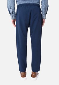 Pantaloni blu navy con cintura elastica, tessuto liscio e vestibilità affusolata, abbinati a scarpe marroni e una camicia di jeans.