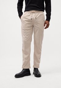 Pantalons en velours côtelé beige avec une taille élastique et un cordon de serrage, présentant des rayures verticales et une coupe décontractée, associés à des bottines noires à la cheville.