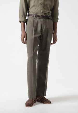 Hombre que lleva camisa de manga larga verde oliva con las mangas remangadas, pantalones a juego, cinturón trenzado marrón y zapatos de ante marrones, de pie frente a fondo blanco.