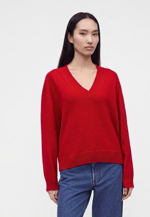 Jeune femme aux longs cheveux noirs raides, portant un pull rouge à col en V et un jean bleu, debout devant un fond clair uni.