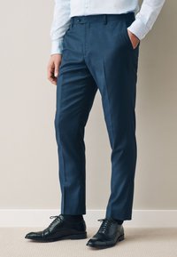 Man wearing tailored navy blue dress pants, light blue shirt, and black leather dress shoes, standing with one hand in pocket.