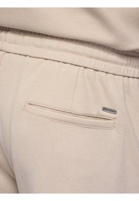 Beige textured fabric pants with elastic waistband, horizontal back pocket, and small metal logo tag above the pocket.