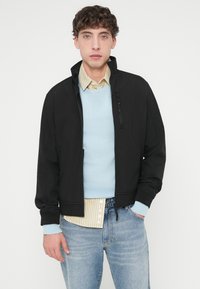 Μαύρο μπουφάν με φερμουάρ και ψηλό γιακά, μαλακό ύφασμα και τσέπη στο στήθος με φερμουάρ. Φορεμένο πάνω από ένα γαλάζιο πουλόβερ και ριγέ πουκάμισο, συνδυασμένο με μπλε τζιν.