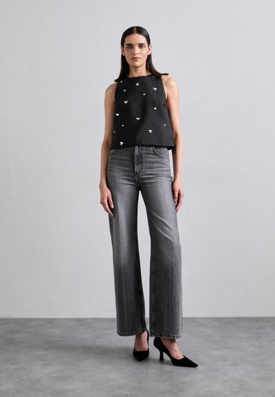 Črna brezrokavnik majica z majhnimi belimi cvetličnimi detajli, v kombinaciji z visokim pasom sivimi širokimi hlačami in črnimi čevlji z oblikovanim nosom.