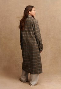 Mujer con cabello largo castaño que lleva un abrigo largo de cuadros marrones y negros sobre pantalones anchos beiges, de pie de lado frente a un fondo beige liso.