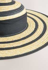 Stråhatt med bred brätt och vävda mönster i beige och svart, utrustad med ett svart tygband runt kronan. Texturerad och naturlig.