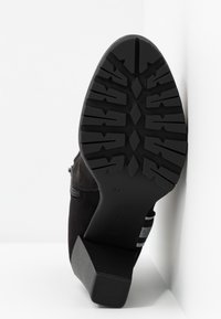 Botín de ante negro que muestra una suela de goma texturizada, tacón cuadrado y un acento a rayas cerca del tacón. Tamaño marcado como 37.