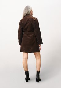 Braunes Cordkleid mit tailliertem Gürtel, langen Ärmeln und kurzem Saum, kombiniert mit schwarzen Ankle Boots mit einem quadratischen Absatz.