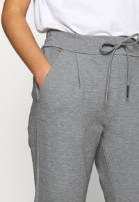 Šedé joggingové kalhoty vyrobené z měkké látky s elastickým pasem na šňůrku, bočními kapsami a zúženým střihem. Hladká textura, minimalizované detaily.