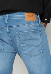 Světle modré džíny z denimu s koženou nášivkou na zadním pásu. Obsahují dvě zadní kapsy s zakřiveným švem.