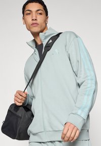 Világoskék cipzáros dzseki puha anyagból, magas gallérral, mindkét ujján három függőleges csíkkal, és egy hozzá illő fekete táskával.