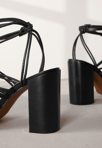 Sorte strop-sandaler med høj hæl, der har et fletdesign og en kraftig, mat sort blokhælt. Glat tekstur og alsidigt design.
