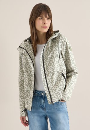 Femme portant une veste à capuche imprimé léopard gris sur une chemise blanche et un jean bleu, debout avec une main dans la poche.