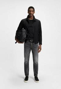 Schwarze Jacke mit Reißverschluss, schwarzes Hemd darunter, graue Jeans und schwarze Sneaker; trägt eine schwarze strukturierte Tasche.