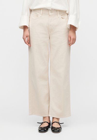 Μπεζ παντελόνι με φαρδύ πόδι από βαμβάκι με διακριτική υφή, με πλαϊνές τσέπες και κούμπωμα με κουμπί, σε συνδυασμό με μαύρα παπούτσια.