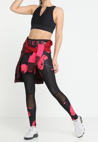 Czarny stanik sportowy z wysokim kołnierzem; czerwony sweter związany w talii; czarne legginsy z kwiatowym wzorem i siateczkowymi akcentami; szare buty sportowe.