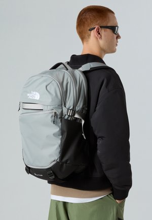 Mochila de nailon gris con acentos negros, múltiples compartimentos y logo. Llevada en el hombro sobre una chaqueta negra y pantalones verde claro.