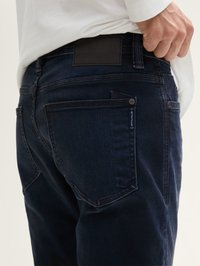 Jeans de denim azul oscuro con un ajuste Slim, que cuentan con bolsillos traseros, un parche de cuero negro en la cintura y detalles de costura sutiles.