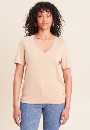 Frau mit lockigem Haar, die ein beiges T-Shirt mit V-Ausschnitt, blaue Jeans mit Blumenstickerei, goldene Creolen und mehrschichtige goldene Halsketten trägt.