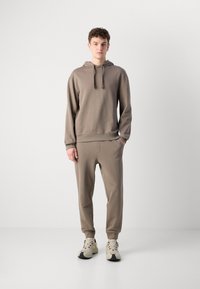 Hanorac maro cu șnururi și pantaloni joggers asortați, ambele dintr-un material moale și neted. Ținuta are manșete cu model canelat și un detaliu subtil cu logo.