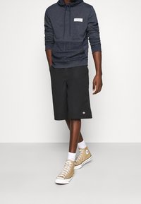 Felpa blu marina con pannello inferiore testurizzato e logo bianco, abbinata a pantaloni corti neri fino al ginocchio e sneakers beige alte.