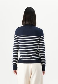 Marineblå genser med hvite horisontale striper, med rund hals og lange ermer. Ribbestrikkede mansjetter og nederkant forbedrer designet.