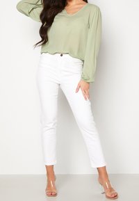 Hellgrüne Bluse mit langen Ärmeln und V-Ausschnitt, kombiniert mit weißen, eng anliegenden Jeans und klaren Sandalen mit Blockabsatz. Glattes Material, lockere Passform.
