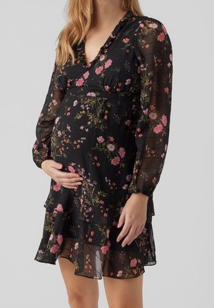 Gravid kvinde iført en sort blomstret langærmet kjole, der holder sin mave med den ene hånd, mod en enkel lys baggrund.