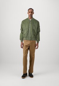 Jachetă bomber verde măsliniu cu două buzunare frontale și mansete din tricot, asortată cu pantaloni bej și pantofi negri cu talpă groasă.