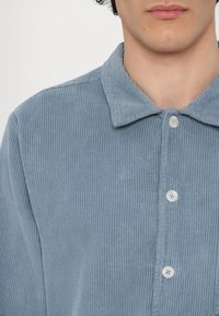 Light blue corduroy shirt with a collar, featuring vertical stripes and white buttons. Texture is soft and slightly ribbed.