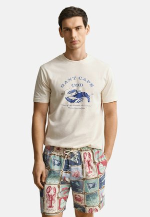Mann trägt ein cremefarbenes T-Shirt mit einer blauen Hummer-Grafik und mehrfarbige Shorts mit nautischen und Hummer-Mustern, steht mit einer Hand in der Tasche.