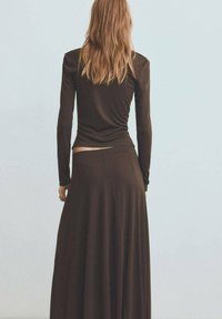 Top lung cu mâneci lungi de culoare maro, purtat cu o fustă maxi fluidă, tot de culoare maro. Materialul pare moale; designul include un corset strâns și o drapează lejeră.