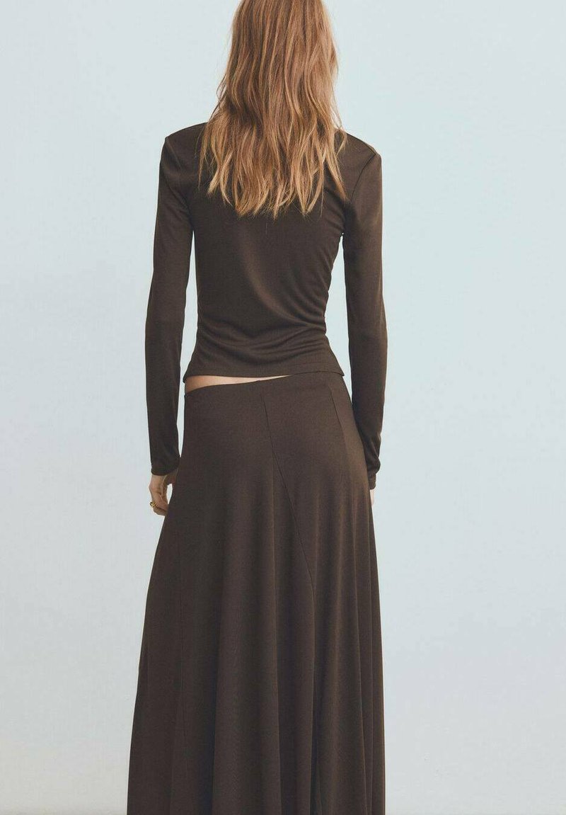 Top lung cu mâneci lungi de culoare maro, purtat cu o fustă maxi fluidă, tot de culoare maro. Materialul pare moale; designul include un corset strâns și o drapează lejeră.