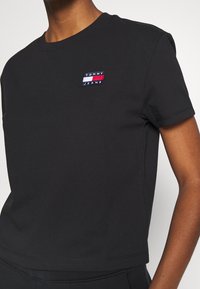 T-shirt in cotone nero con maniche corte e collo rotondo. Presenta un logo ricamato di Tommy Jeans in rosso, bianco e blu sul petto.