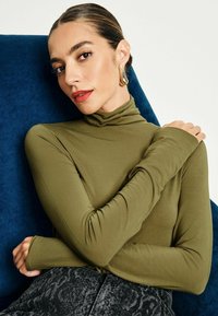 Olivgrön långärmad polotröja med en tight design och ribbad textur. Matchar med mörka, mönstrade högmidjade byxor.