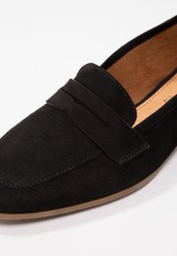 Mocassins en suède noir avec une semelle en caoutchouc plate et une sangle décorative en cuir noir sur le devant. Intérieur doublé d'un matériau beige clair.