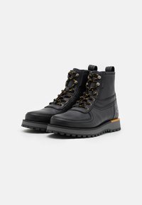 Black leather boots with metallic eyelets, golden laces, padded collar, textured sole, and smooth finish. Reinforced heel and logo detail.