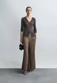 Bruine cardigan met knopen, gecombineerd met lichtbruine wijde broekspijpen. Model houdt een kleine zwarte handtas vast, staand op een grijze vloer.