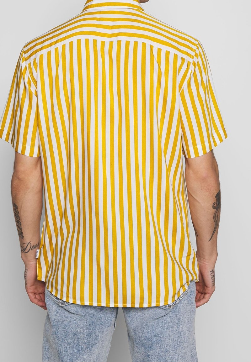 Personne portant une chemise à manches courtes rayée verticalement jaune et blanche et un jean bleu clair, vue de dos.