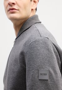 Giacca bomber grigia con collo a costine texturizzato, tessuto grigio chiaro e un patch quadrato grigio sul maniche sinistro.