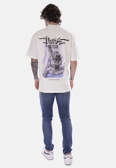 T-shirt bianco con una grafica sulla schiena raffigurante un teschio con un serpente, accentuata da lettere nere e dettagli viola, abbinata a jeans blu.