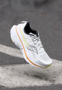 Weißer Sportschuh mit Obermaterial aus Mesh, kontrastierendem schwarzen Innenfutter, gelben und orangefarbenen Akzenten, gepolsterter Sohle und auffälliger Markenkennung an der Seite.