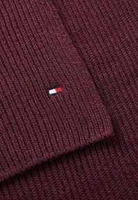 Bordeaux ribbet strikstof med et lille broderet logo i rød, hvid og blå i et hjørne. Blød tekstur med synlige syninger.